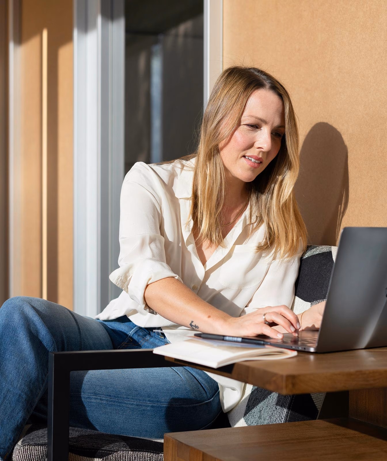 Woman with blonde hair in a white shirt and jeans working on a laptop at a wooden table with a notebook and a white water bottle.